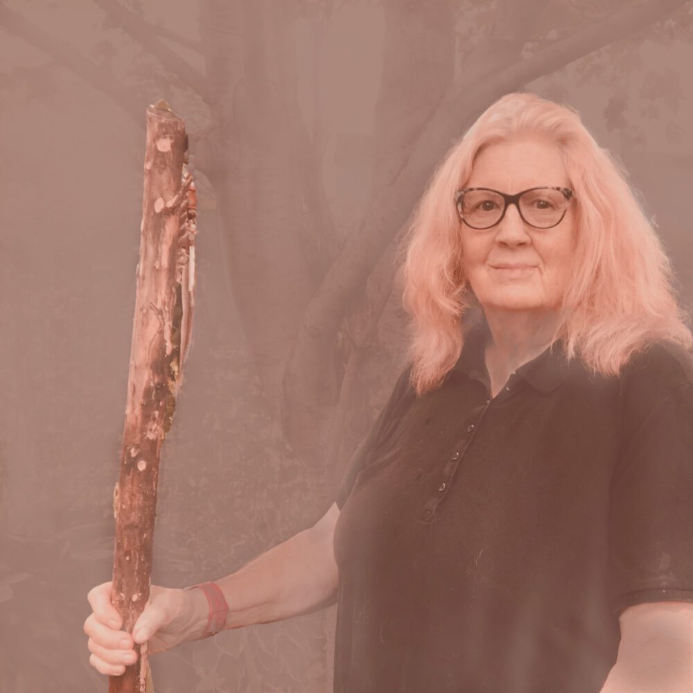 A woman with long hair standing, holding a tree-branch stake in the ground.